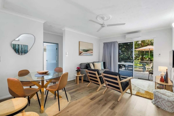 A modern living room with a glass dining table, wooden chairs, a sofa, and a ceiling fan.