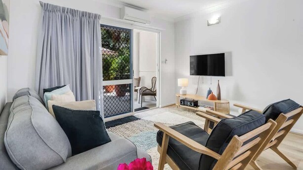 A modern living room with a sofa, chairs, a TV, and a sliding glass door leading to an outdoor area. - Leafy Solace—A Contemporary Cairns City Hideaway (Cairns)