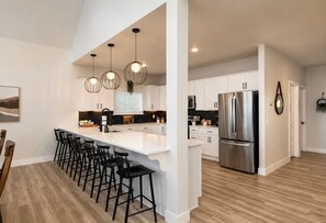 Een moderne keuken met een centraal eiland, barkrukken, roestvrijstalen apparatuur, hanglampen en uitzicht op de eetruimte.