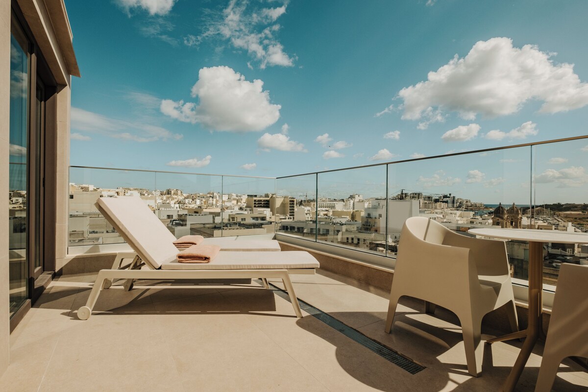 Une terrasse sur le toit, dotée de chaises longues et d’une table, offrant une vue sur la ville.