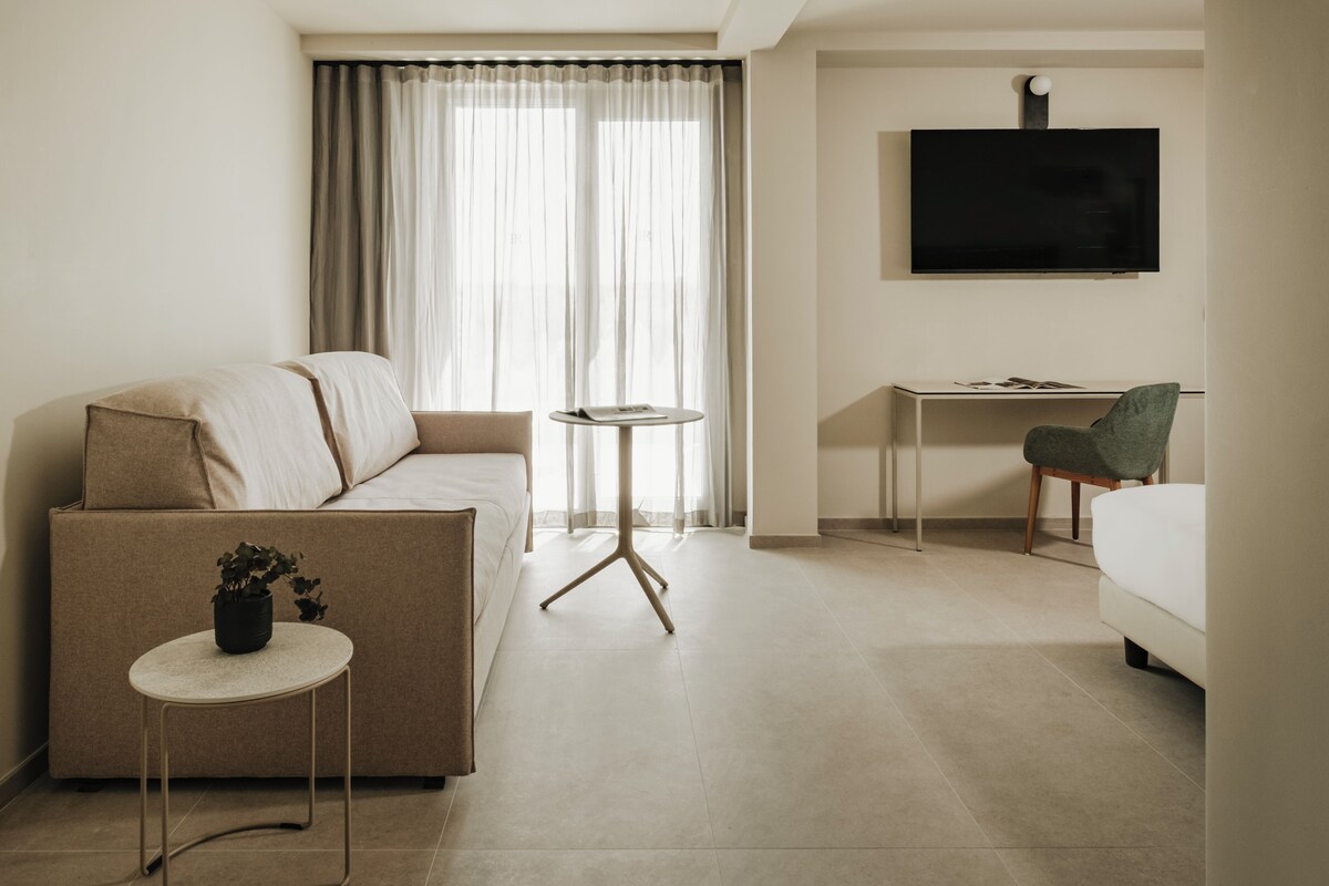 Une chambre d’hôtel moderne, équipée d’un canapé, d’une petite table ronde, d’un bureau et d’un téléviseur fixé au mur.