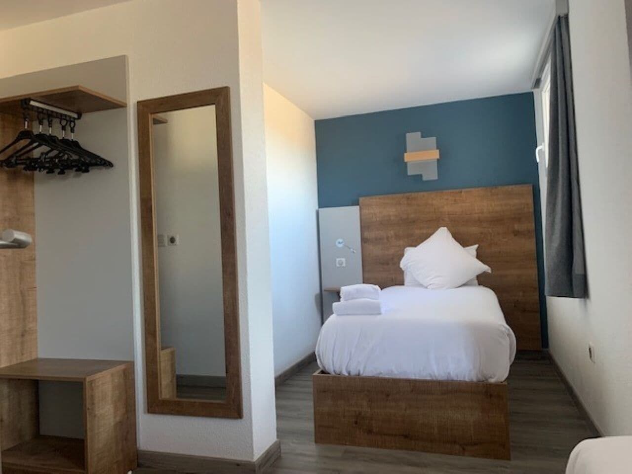 Una habitación de hotel con cama, cabecera de madera, espejo, banco de madera, toallero en la pared y ventana con cortinas.