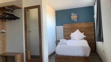 Una habitación de hotel con cama, cabecera de madera, espejo, banco de madera, toallero en la pared y ventana con cortinas.
