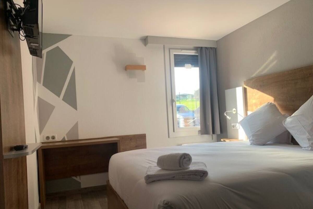 Una habitación de hotel con una cama, cabecera de madera, mesita de noche, ventana con cortinas y vistas al exterior.