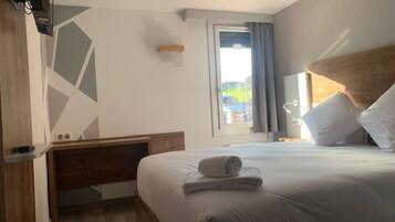 Una habitación de hotel con una cama, cabecera de madera, mesita de noche, ventana con cortinas y vistas al exterior.