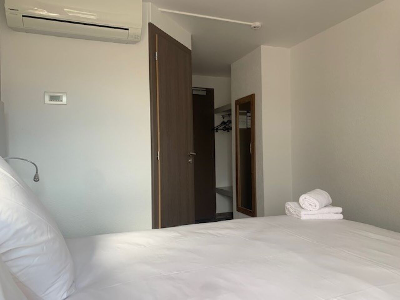 A hotel room with a bed, an air conditioning unit, a door, and a mirror.