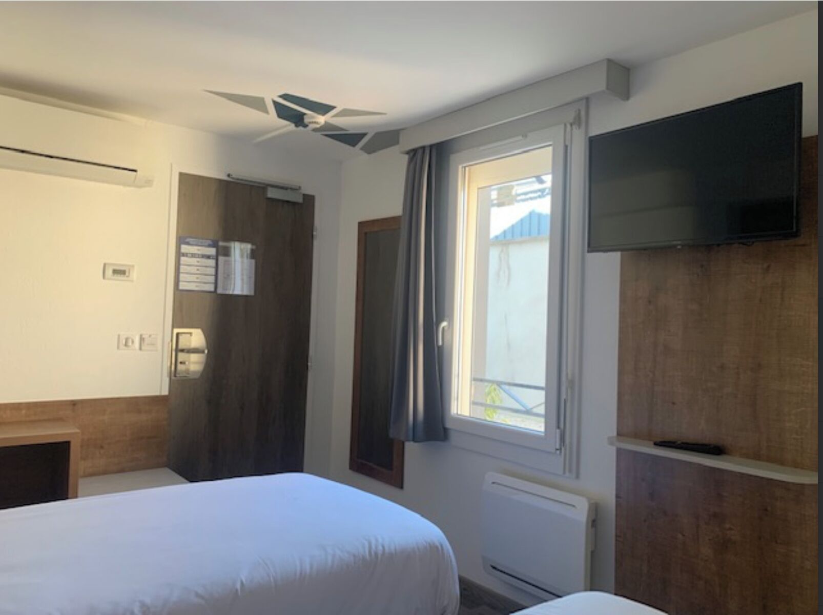 Habitación de hotel con dos camas, ventilador de techo, televisión, ventana con cortinas y radiador.