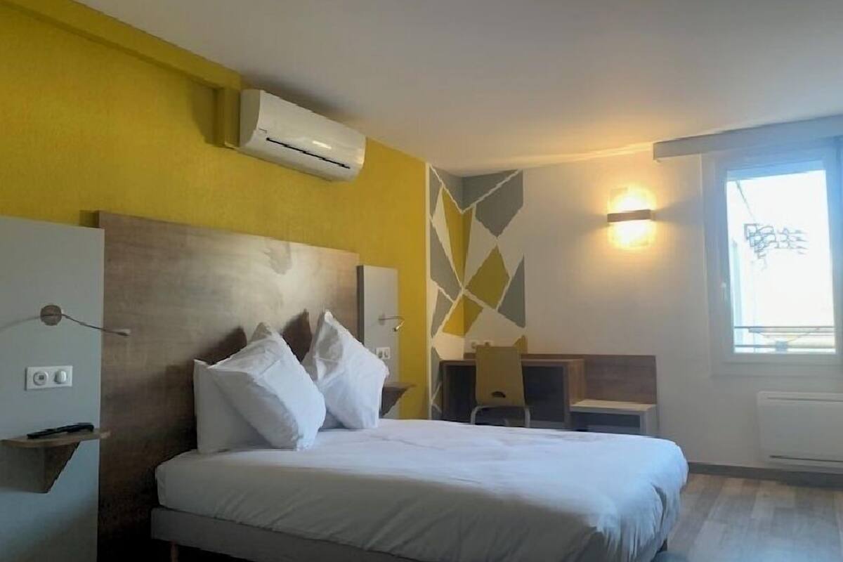 Habitación de hotel con cama, escritorio y silla. Hay un aire acondicionado en la pared y una ventana con vistas.