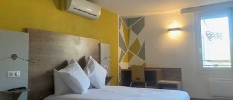 Habitación de hotel con cama, escritorio y silla. Hay un aire acondicionado en la pared y una ventana con vistas.