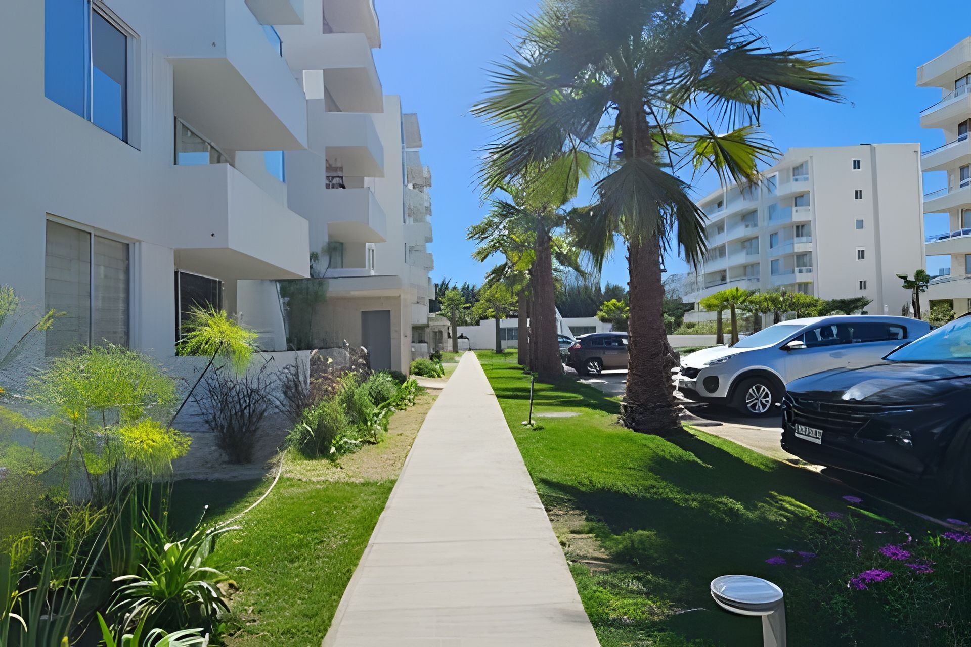 Un moderno edificio residencial con un camino central, coches aparcados y abundante vegetación.