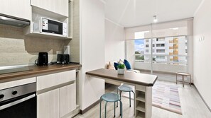A modern kitchen with a microwave, toaster, and coffee maker. There is a dining area with a table and two stools. The room has a large window with a view of buildings. - Oceana Suites Distrito Verde (La Serena)