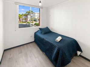 A single bed with a blue quilt and a rolled towel, a window with a view of palm trees and buildings, and a light fixture above the bed. - Oceana Suites Distrito Verde (La Serena)
