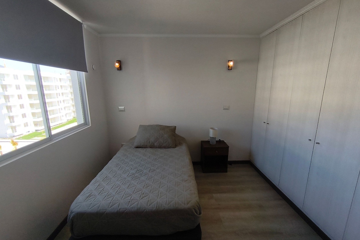 A bedroom with a bed, a nightstand, a lamp, a wardrobe, and a window with a view of buildings.