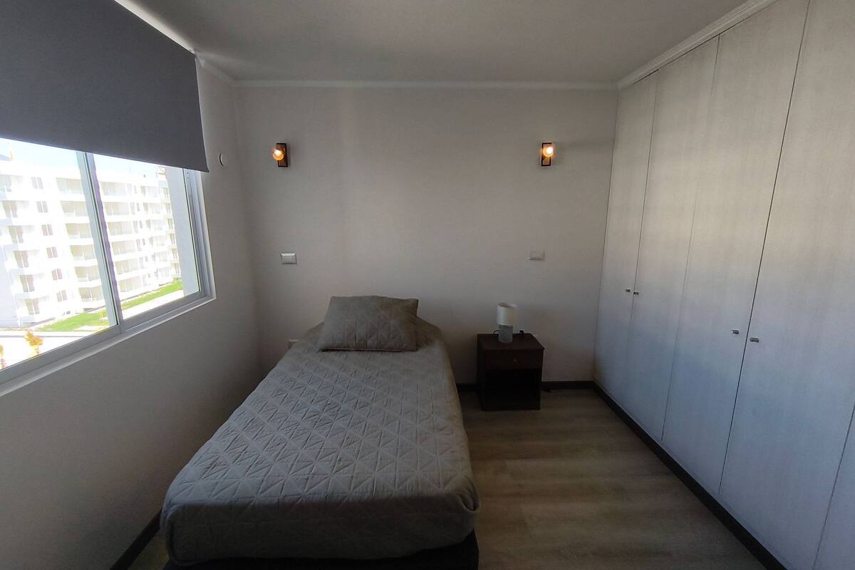 A bedroom with a bed, a nightstand, a lamp, a wardrobe, and a window with a view of buildings.