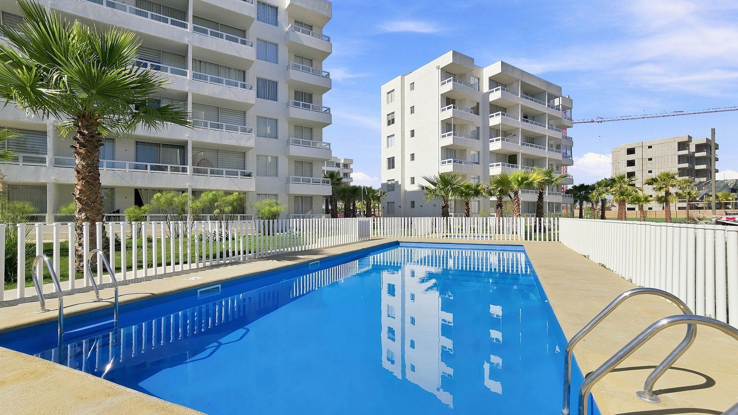 Una piscina con valla blanca, rodeada de palmeras y un edificio de varios pisos con balcones.