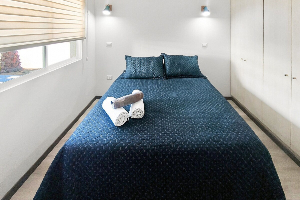 Quarto de hotel com cama, duas almofadas, janela com persianas e toalha sobre a cama.