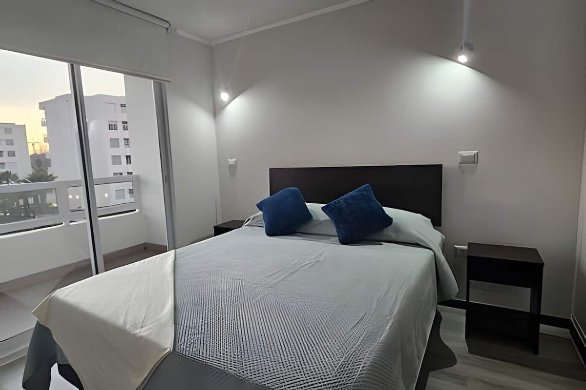 A modern bedroom with a large bed, two pillows, a nightstand, and a view of buildings through a large window.