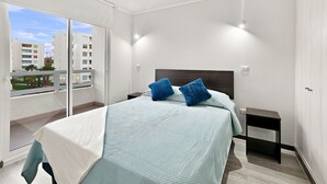 A bedroom with a bed, two pillows, a bedside table, and a view of buildings through a sliding door. - Oceana Suites Distrito Verde (La Serena)