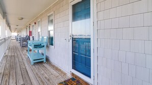 Une terrasse en bois couverte, avec une rangée de chaises et de tables bleu clair, une porte bleue et un mur extérieur blanc.