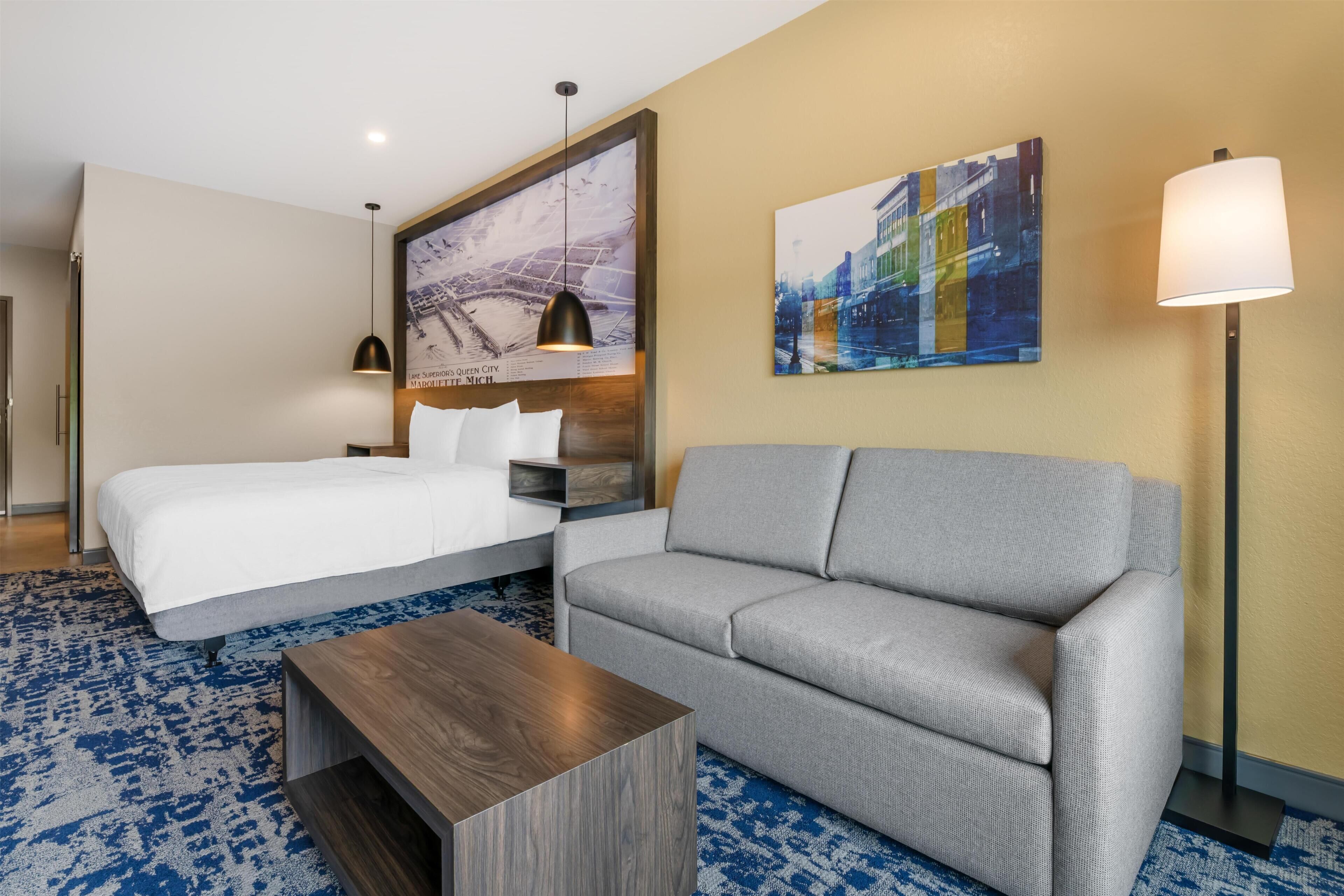 A hotel room with a bed, a gray sofa, a wooden coffee table, a floor lamp, and two wall art pieces.