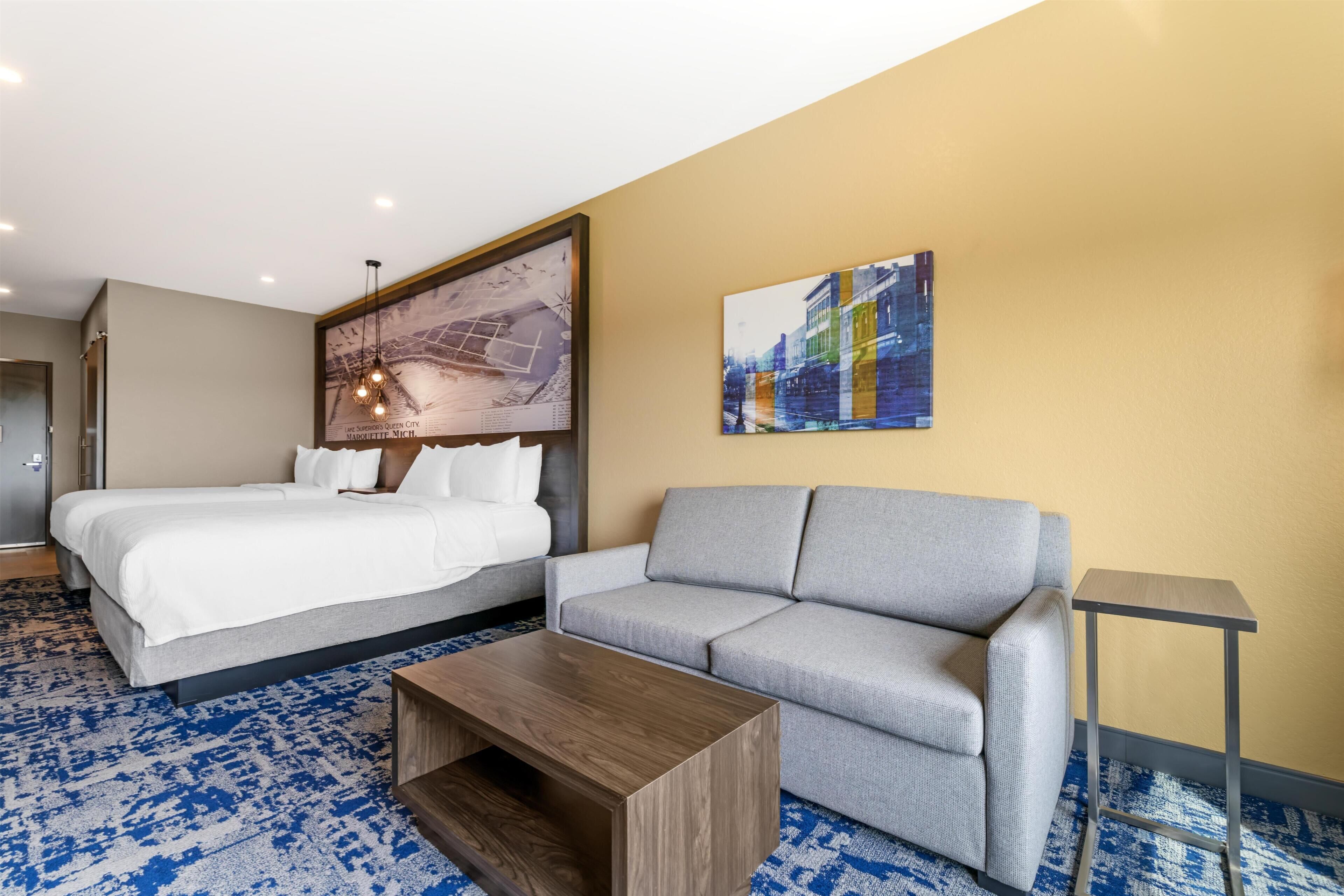 A hotel room with a bed, a gray sofa, a wooden coffee table, a framed cityscape artwork, and a blue patterned carpet.