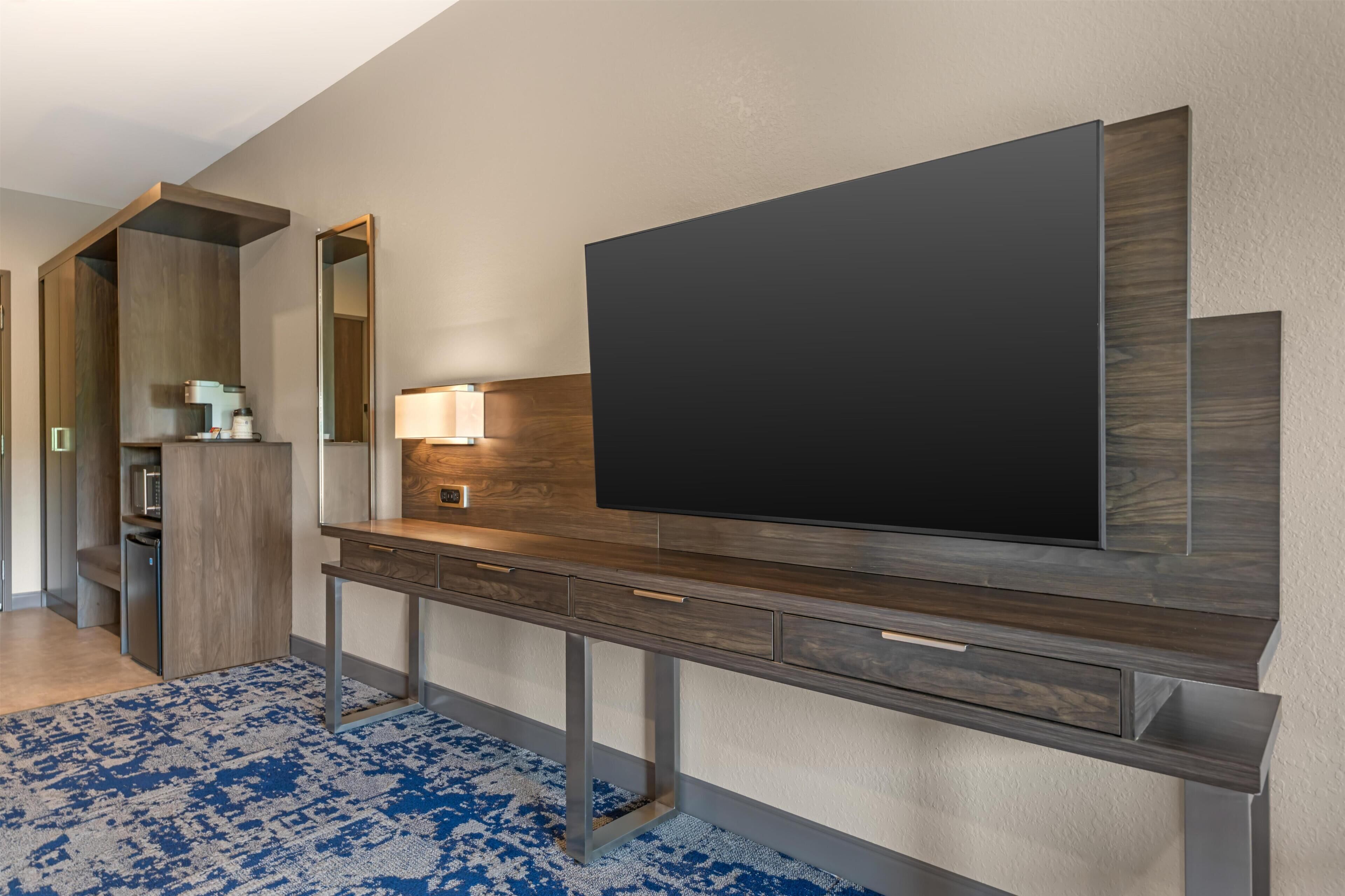 Una habitación de hotel moderna con un televisor de pantalla plana grande montado sobre una consola de madera, un escritorio empotrado con cajones, un espejo y una alfombra azul estampada.
