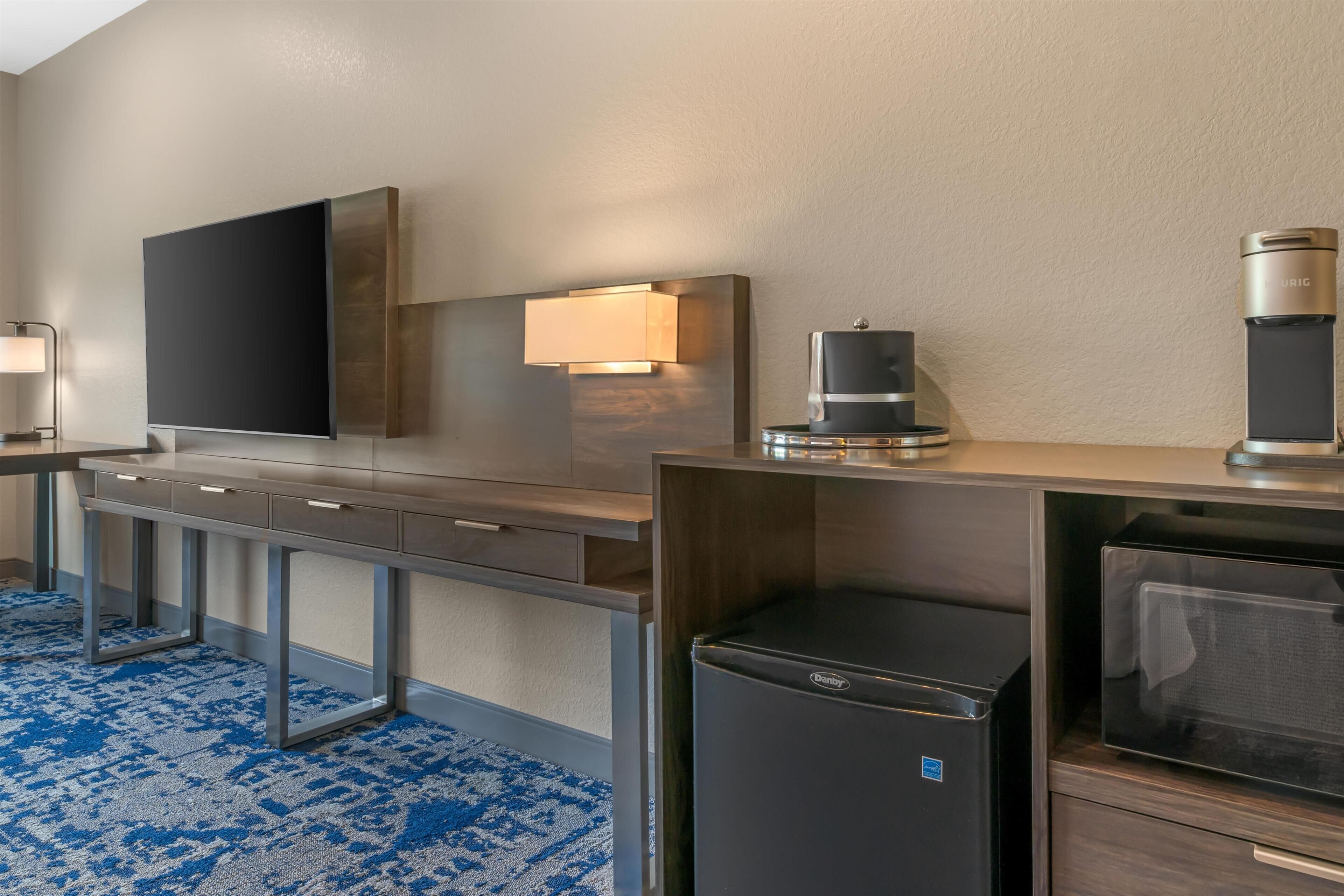 Habitación de hotel con televisión de pantalla plana, cafetera, refrigerador, microondas y escritorio con cajones.
