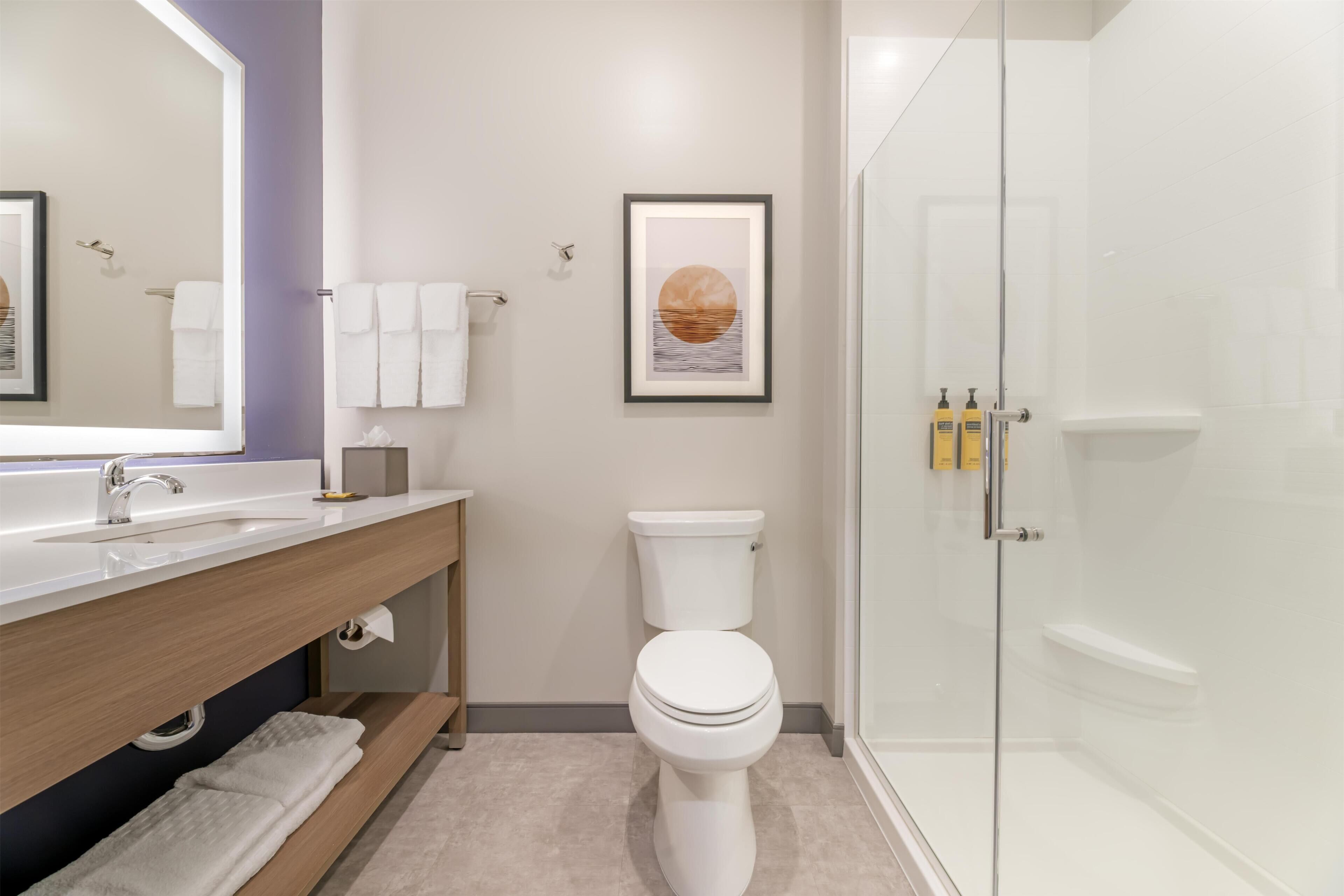 A modern bathroom with a white sink, toilet, and shower. There is a framed picture on the wall and toiletries on the sink.