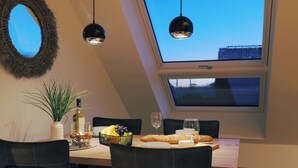 Un coin repas avec une table dressée pour deux personnes, une plante en pot, une bouteille de vin et un saladier de fruits. Deux lampes suspendues noires sont placées au-dessus de la table, et une fenêtre offre une vue sur le ciel.