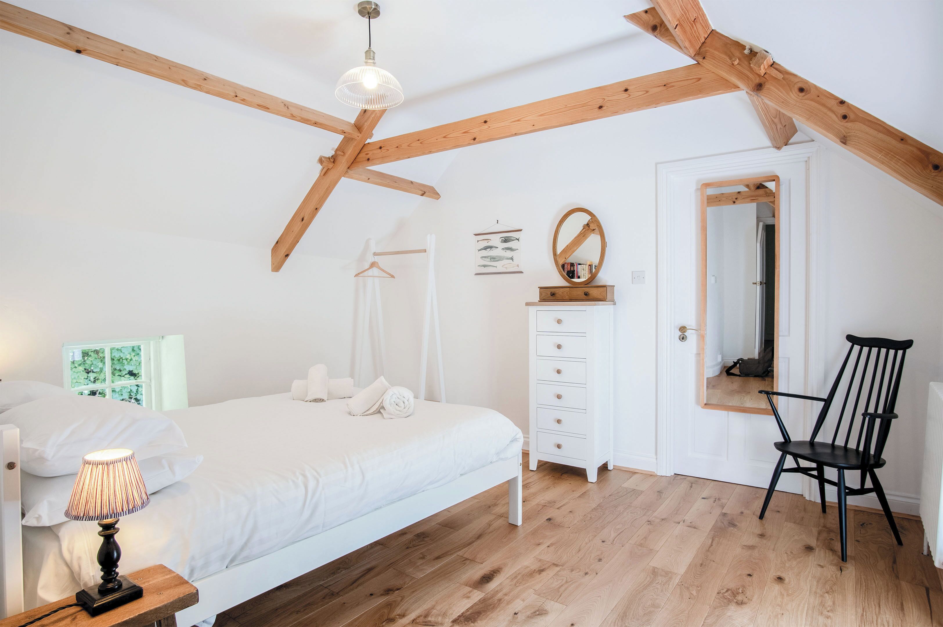 A bedroom with a wooden ceiling, a bed with white bedding, a bedside table with a lamp, a dresser, a mirror, and a chair.