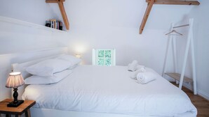 Ein Schlafzimmer mit Holzbalkendecke, einem Bett mit weißer Bettwäsche, einem Nachttisch mit Lampe, einem Kleiderständer und einem gerahmten Bild an der Wand.