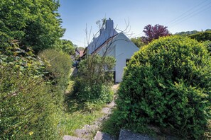 A white building with a steep roof, surrounded by dense greenery and a stone pathway leading to the entrance. - Briardene - 2 Bedroom Cottage - Oxwich (Swansea)