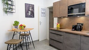 Une cuisine compacte dotée d’un plan de travail en bois, d’un four à micro-ondes encastré et d’une machine à café. On y trouve un petit coin repas avec des tabourets et une table dressée avec des fruits et une plante en pot.