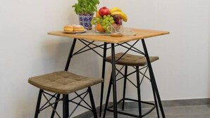 Un petit coin repas avec une table en bois, deux tabourets noirs, une plante en pot et une œuvre d’art encadrée accrochée au mur.