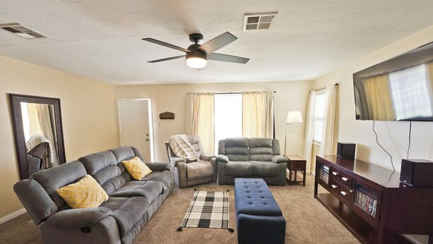 Olohuoneessa on harmaa kulmasohva, sininen jalkarah, nojatuoli, sohvapöytä ja TV-taso, jonka päällä on kirjoja.