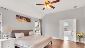 Ein Schlafzimmer mit Bett, Nachttisch, Lampe, Deckenventilator, Kunstwerk und Blick ins Bad.