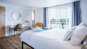 A hotel room with a bed, a desk, a chair, a round mirror, and a view of a body of water through large windows. - Skyline Duplex Penthouse - Llanelli (Llanelli)