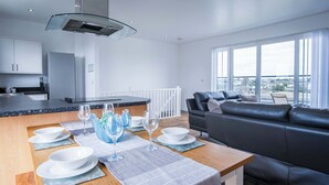 A modern kitchen with a dining area, a black leather sofa, and large windows offering a city view. - Skyline Duplex Penthouse - Llanelli (Llanelli)