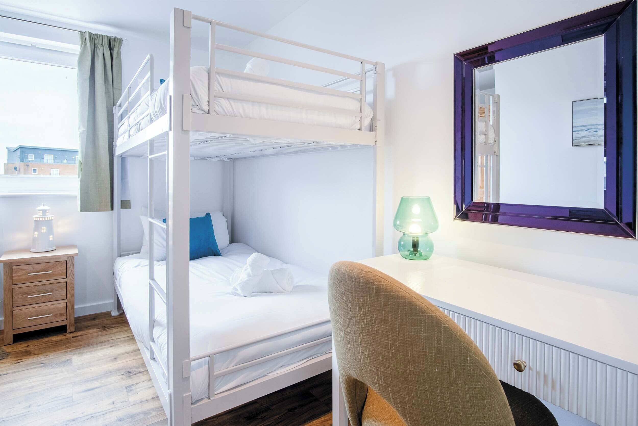 Une chambre avec un lit superposé, un bureau et une chaise, un miroir et une fenêtre avec des rideaux.