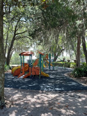 Explore a shaded playground surrounded by mature trees, featuring slides and climbing frames on a textured surface.