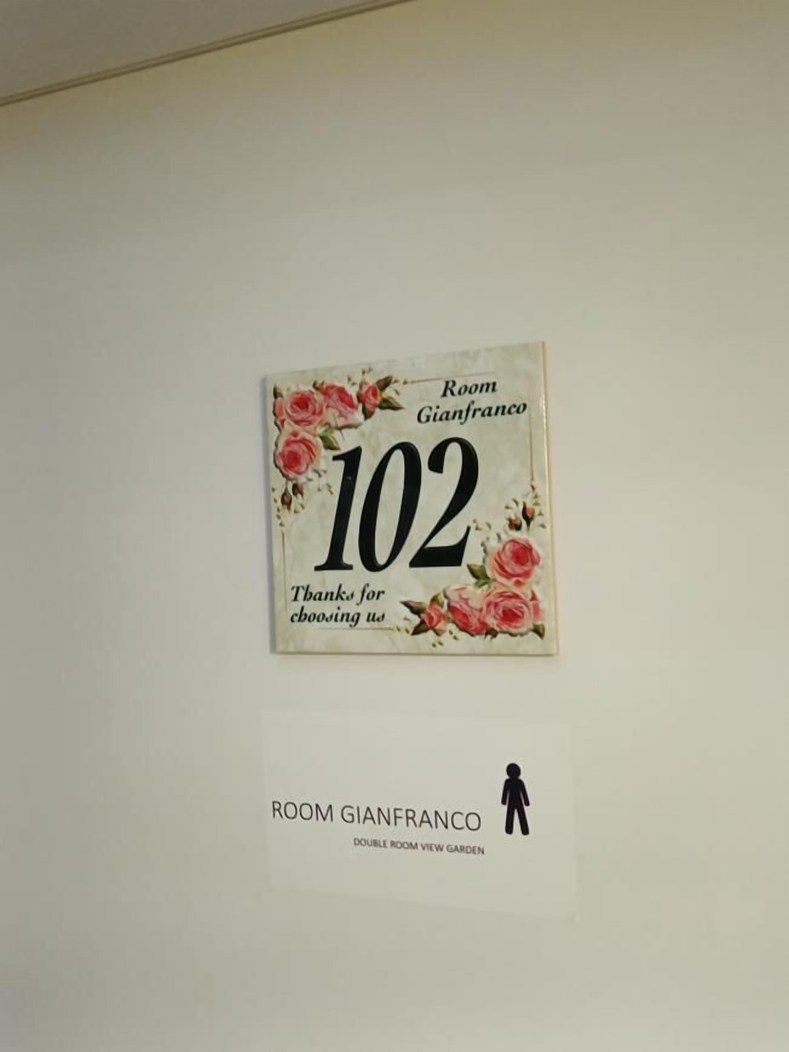 Señal para la Habitación Gianfranco, número 102, con un diseño floral y la silueta de una persona.