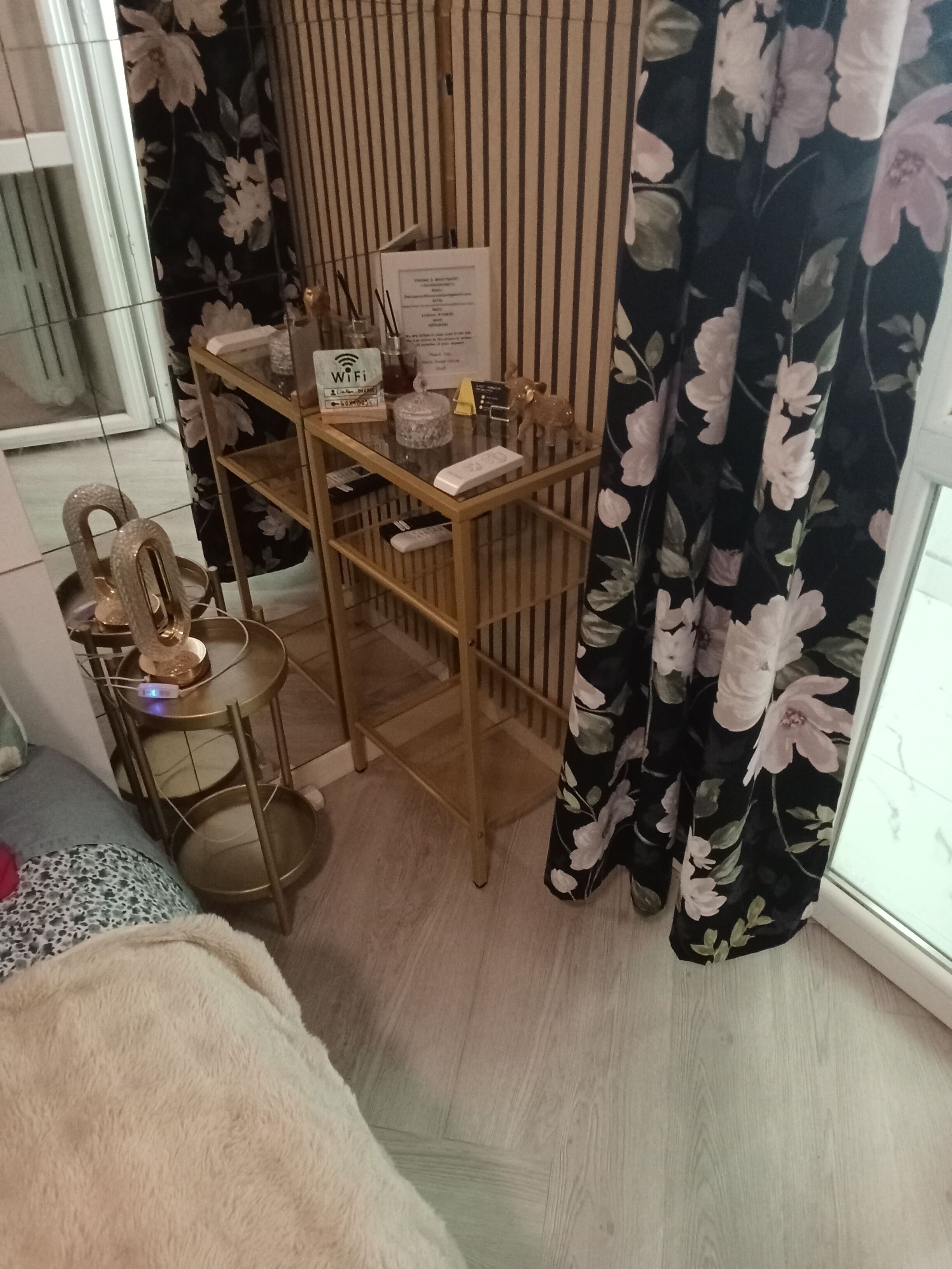 A room with a wooden desk, a chair, a small table with a decorative item, a curtain with a floral pattern, and a window.