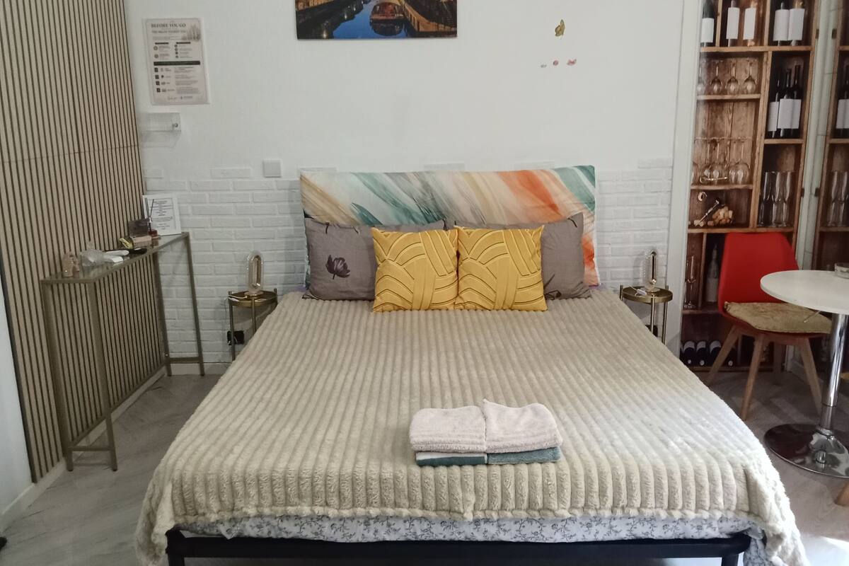 A neatly arranged bedroom with a bed, a bedside table, a chair, a bookshelf, and a wall-mounted picture.
