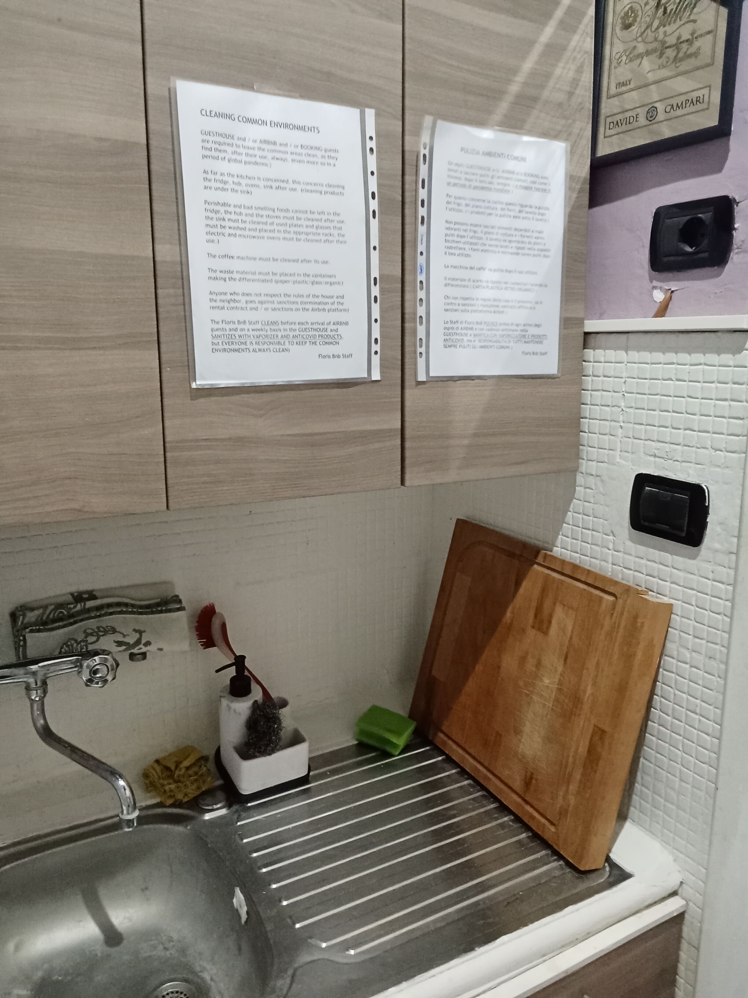 A kitchen with a sink, a wooden cutting board, a green sponge, and a small vase with a flower.