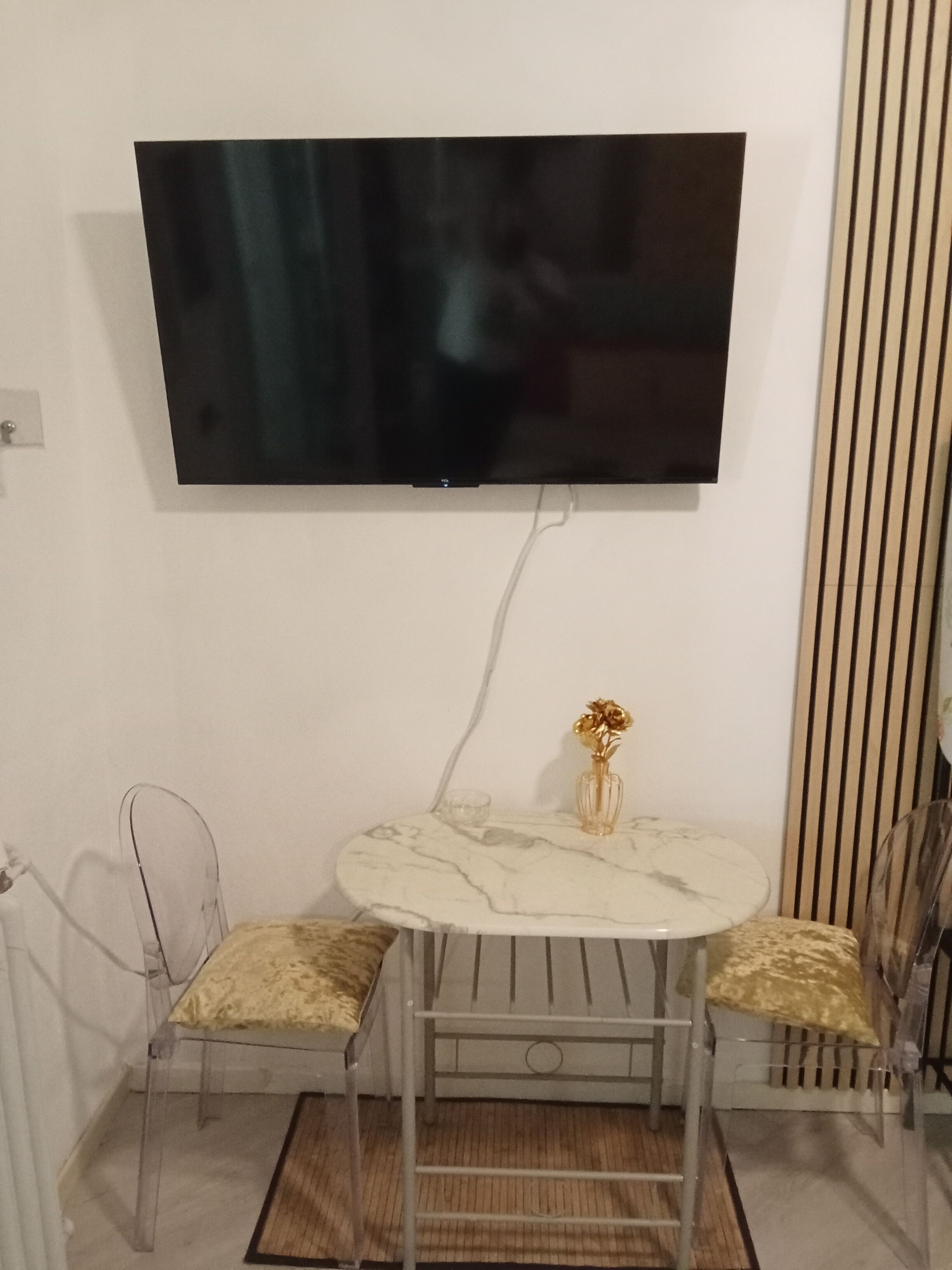 Ein Zimmer mit an der Wand montiertem Fernseher, einem runden Tisch mit Stühlen, einer kleinen Vase und einem Heizkörper.