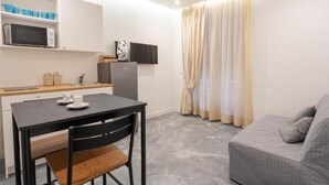 A compact living space with a kitchenette, a dining table with chairs, a grey sofa, and a TV mounted on the wall. - La Maison di Tina (Naples)