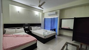A hotel room with two beds, a ceiling fan, an air conditioning unit, a window with blue curtains, and a mirror on the wall. - Hotel Aman International (Kalapara)