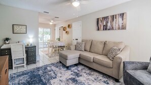 Un salon comprenant un canapé d’angle beige, un fauteuil blanc, un bureau avec une chaise et un ventilateur de plafond.