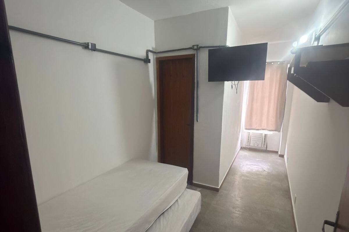 Ein schmaler Flur mit einem Einzelbett links, einer Tür am Ende und einem an der Wand befestigten Fernseher.