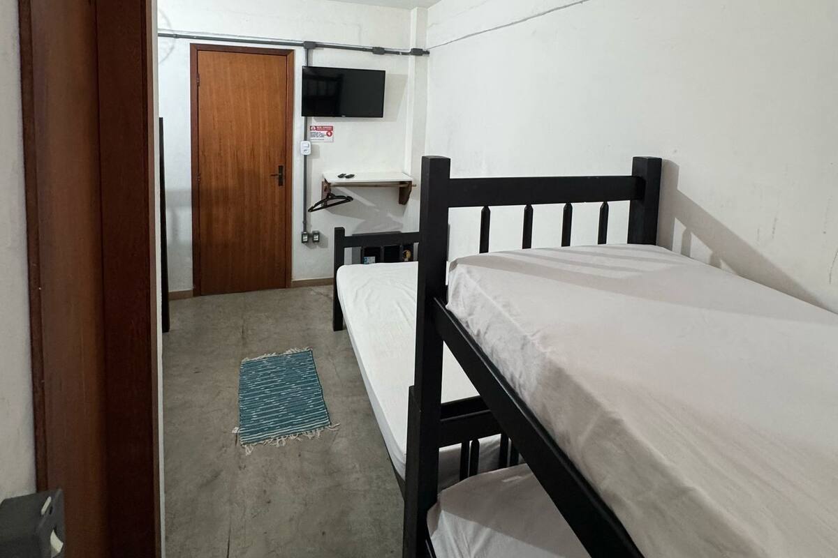 Eine kleine, einfache Unterkunft mit einem hölzernen Bett, einem an der Wand befestigten Fernseher und einer Tür zu einem weiteren Raum.